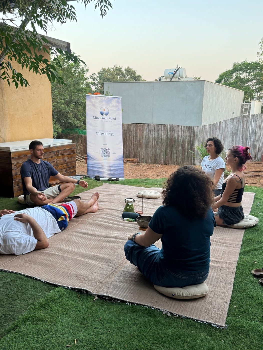 תומר מנחה מפגש מדיטציה קבוצתי במרחב חיצוני לצד שלט Mind Your Mind